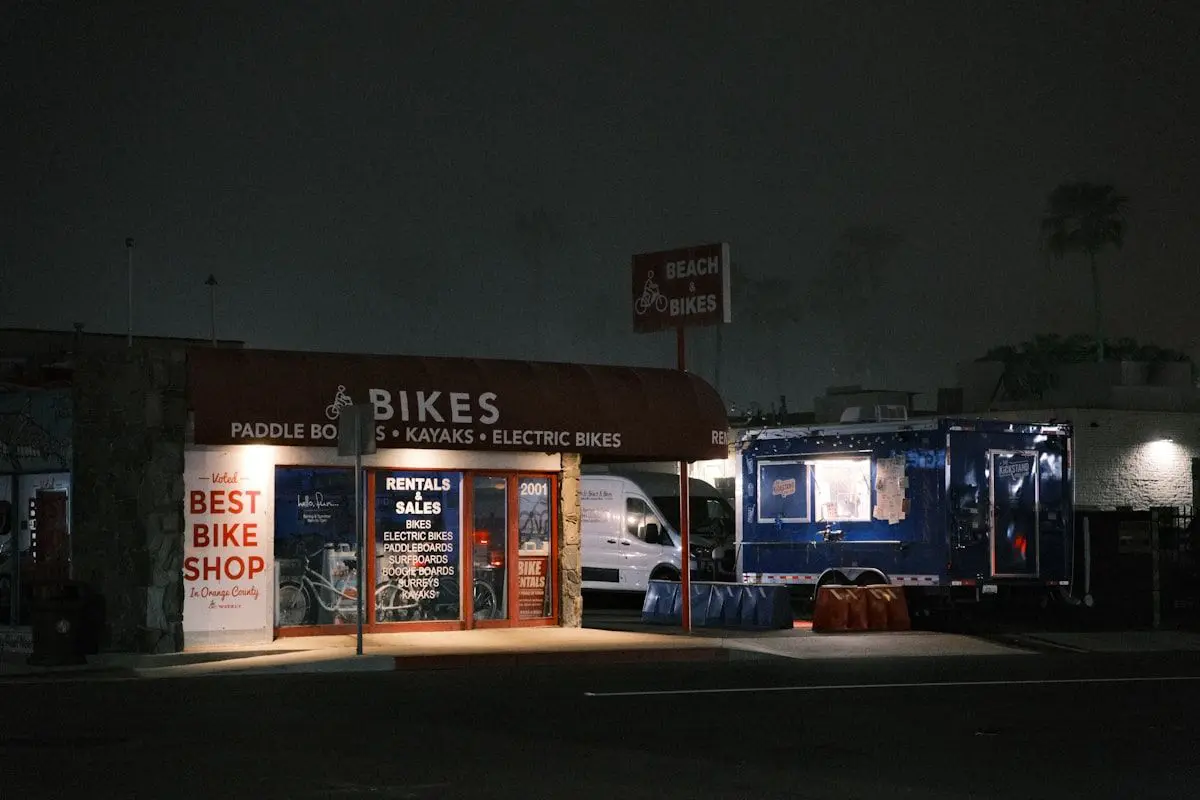 Several rental e-bikes lined up outside a shop with step-through frames, front baskets, rear racks, and visible chargers nearby, with one bike being checked for battery level and another fitted with a helmet