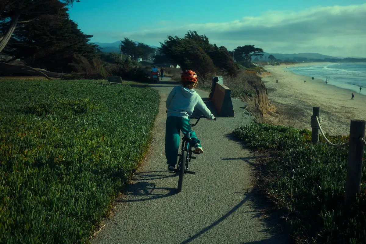 A long two-seat tandem bicycle parked on a sunny coastal path beside a single cruiser and a trike, with helmets and a bike lock resting nearby, showing the different rental options in a flat beachside setting