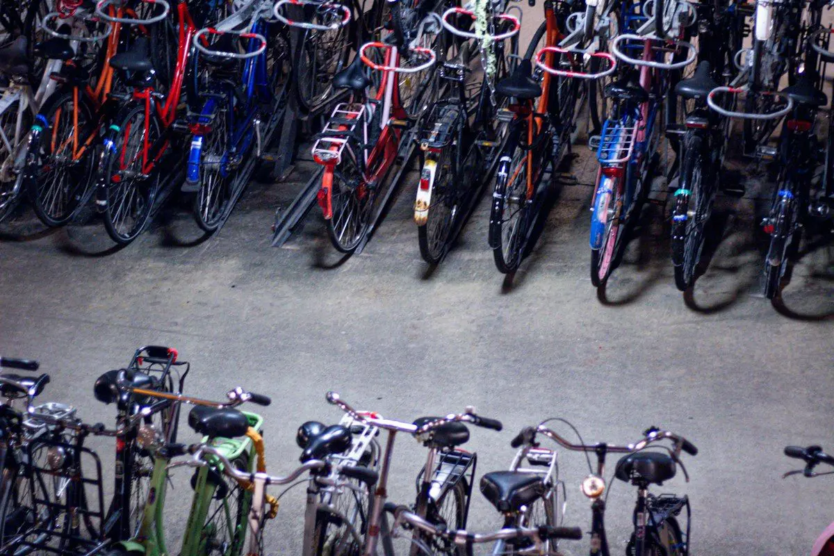 A rental shop floor with several adult bikes arranged for comparison: a road bike, a comfort bike, a mountain bike, and an e-bike, plus a measuring tape laid on the ground and a person’s inseam being measured against a wall with a hardcover book used as a guide, showing the fit process before choosing a bike.