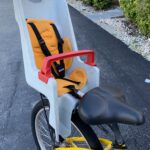 This image shows a child's safety seat mounted on the rear of a bicycle. The seat itself is made of a light gray molded plastic and features bright orange padding on the back and seat area. For safety, it's equipped with a black harness system, including shoulder straps, and a red handlebar-like safety bar in the front.